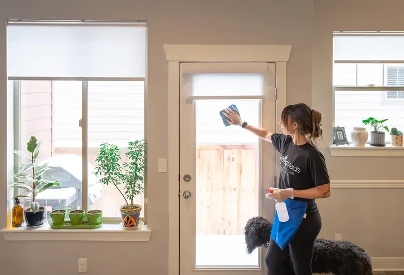 A Dashing Maids cleaner wipes the glass door of a bright, freshly cleaned Denver home while the family dog looks on